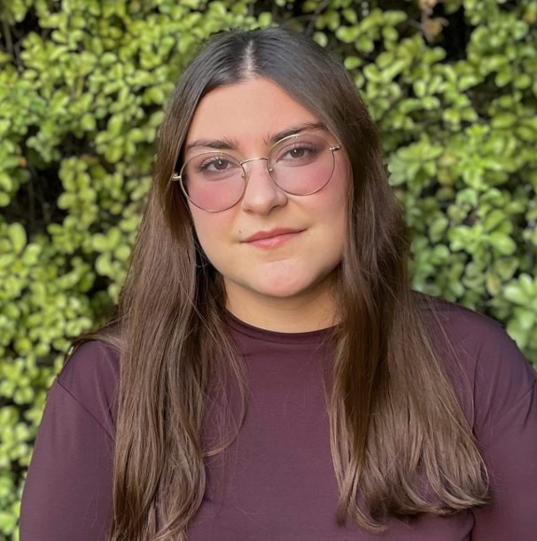 Portrait of a young woman with round glasses and long brown hair wearing a maroon top, standing in front of a green hedge.